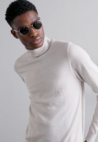 Pull à col roulé gris clair en matériau doux et fin. Dispose d'un design ajusté et d'une texture côtelée au niveau du col. Porté avec des lunettes de soleil sombres.