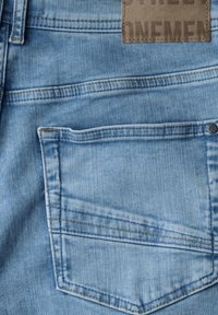 Jeans de denim azul claro con un solo bolsillo trasero y una etiqueta de cuero marrón en relieve con texto. Textura suave y diseño casual.