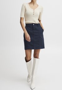 Pull en ivoire à côtes avec des manches courtes et des détails de boutons, associé à une jupe ajustée marine avec poches et une fermeture à boutons à l'avant. Bottes blanches jusqu'aux genoux.