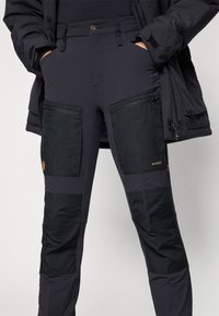 Zwarte outdoorbroek met een combinatie van gladde en gestructureerde stof, voorzien van ritszakken en versterkte panelen in contrasterend zwart.