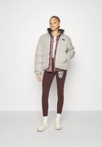 Šedá zimní bunda s černými akcenty, vínová košile na knoflíky, hnědé legíny s bílým logem Nike a bílé ponožky s žlutými teniskami.