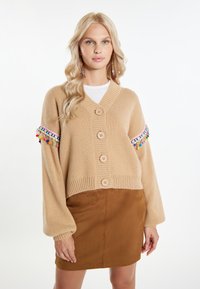 Cardigan beige lavorato a maglia con cinque bottoni, polsini a coste e accenti in frange colorate sulle maniche, abbinato a una mini gonna in suede marrone.