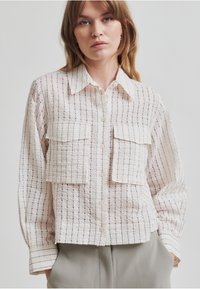 Camisa abotonada ligera y texturizada en color crema con finas rayas verticales y dos bolsillos frontales. Cuenta con un collar clásico y mangas largas.