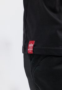 Must musta, millel on sile tekstuur, esiküljel punane silt "ALPHA INDUSTRIES INC. U.S.A." alumises vasakus nurgas. Lihtne disain.