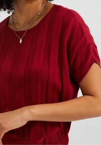 Červený pletený top s vertikálním žebrováním, krátkými rukávy a kulatým výstřihem. K dispozici jsou vrstvené stříbrné řetízky jako doplňky.