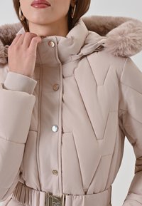 Vrouw in een lichtbeige gewatteerde winterjas met een met bont omzoomde capuchon, drukknoopjes en een riem met een gouden gesp, die de kraag aanpast.