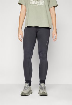 Zwarte legging met een nauwsluitend ontwerp, gladde textuur en zichtbare stiknaden. Gepaard met een losse olijfgroen T-shirt en grijze sportieve schoenen.