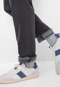 Hvite sneakers med blå og grå detaljer, båret med mørke bukser rullet oppe ved anklerne, som fremviser teksturerede grå sokker med mønstrede manchetter.