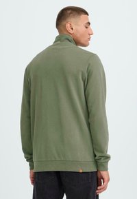 Olivgrüner Pullover mit hohem Kragen, aus weichem Stoff gefertigt. Verfügt über gerippte Bündchen und ein Etikett-Detail am Saum. Schlichte und lockere Passform.