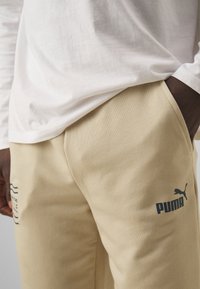 Bež hlače iz frotirja z mehkim otipom, z logotipom PUMA v temno modri barvi in stranskimi žepi. Nosi se z belim dolgim rokavom.