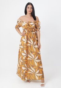 Abito maxi off-the-shoulder in marrone terroso con grandi motivi floreali gialli e bianchi. Presenta una vita arricciata e maniche svasate.