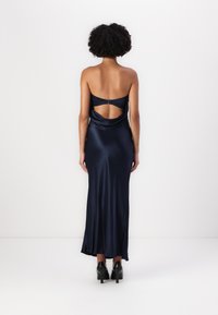 Ναυτικό σατέν φόρεμα με ανοιχτή πλάτη και κοψίματα, διαθέτοντας εφαρμοστή σιλουέτα και μήκος μέχρι το γόνατο, συνδυασμένο με μαύρες ψηλές γόβες.