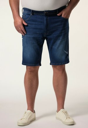 Mann trägt dunkelblaue Jeansshorts und weiße Sneaker, steht mit den Händen nahe den Taschen vor einem einfarbigen Hintergrund.