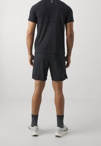 Černé sportovní tričko s krátkým rukávem a zaobleným lemem, spárované s černými šortkami. Na nohou má šedé vzorované ponožky a bílé tenisky.