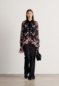 Blouse noire transparente avec motif floral, manches longues, col noué ; associée à un pantalon noir à rayures et un petit sac noir.