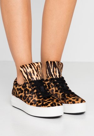 Jambes portant des baskets à plateforme à imprimé léopard avec des lacets noirs sur une surface et un arrière-plan gris clair.