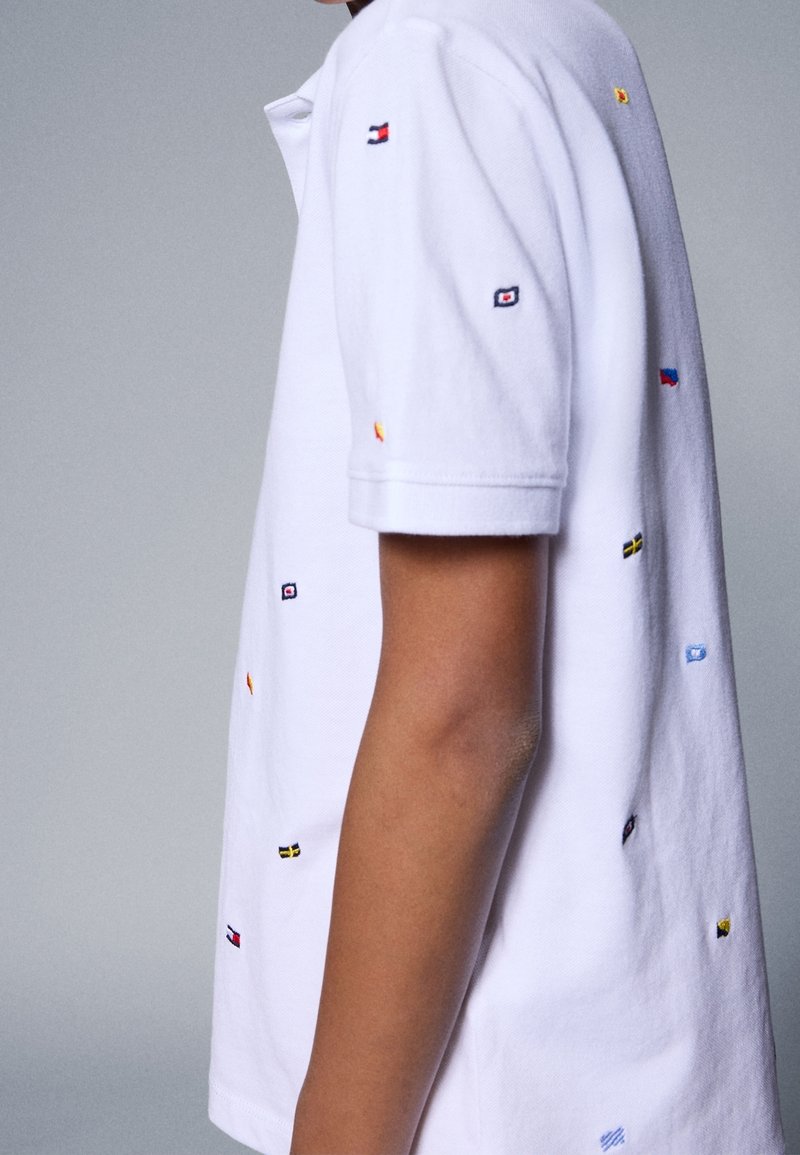 Personne portant une chemise blanche à manches courtes avec de petits drapeaux brodés multicolores dispersés sur le tissu sur un fond uni.