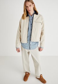Giacca cropped beige con colletto nero, indossata sopra una camicia a righe blu. Pantaloni larghi di colore chiaro e sneaker marroni completano l'outfit.