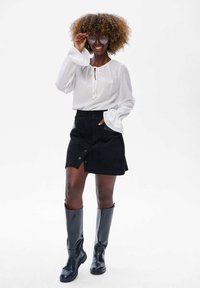 Witte blouse met klokmouwen, zwarte suède mini-jurk met knoopdetails en zwarte kniehoge laarzen. Model poseert met zonnebril.