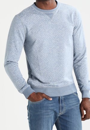 Mann trägt hellblauen gemusterten Langarm-Sweatshirt mit einer Hand in der Tasche der blauen Jeans vor weißem Hintergrund.