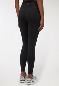 Czarne legginsy z wysokim stanem o gładkiej teksturze i zaokrąglonych szwach. W zestawieniu z szarymi i granatowymi butami sportowymi.