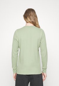 T-shirt à manches longues à côtes dans un vert clair, avec un col rond et des manches ajustées. Le tissu présente un motif gaufré texturé.