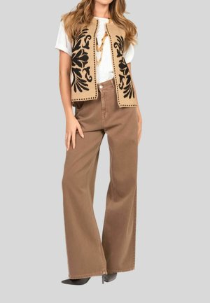 Gilet beige ricamato con design floreale nero sopra una camicia bianca, abbinato a pantaloni larghi marroni a vita alta. Tacchi neri completano il look.