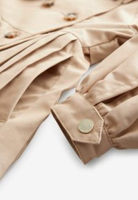 Beige jakke med plisseret design, dekoreret med skinnende guldknapper og et glat, struktureret stof. Rene, simple linjer og skræddersyet pasform.