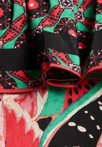 Tessuto colorato con motivi di farfalle rosse e nere su uno sfondo verde, con elementi floreali e un bordo piegato.