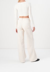 Vit ribbad långärmad crop top i kombination med ljusbeige vida byxor, med bakfickor och en texturerad yta. Svarta platta skor synliga.