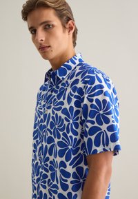 Camisa de manga corta de algodón blanco con un estampado floral azul. Presenta un cierre de botones en la parte frontal y un diseño de cuello clásico.