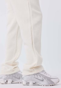 Piernas inferiores con pantalones holgados color crema y zapatillas Nike blancas y plateadas con suelas de amortiguación de aire visibles sobre un fondo blanco.