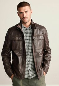 Braune Lederjacke mit zwei Brusttaschen, Druckknopfverschlüssen und glatter Oberfläche, getragen über einem schwarz-weißen karierten Hemd.