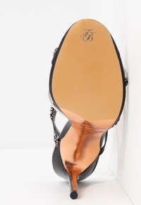 Chaussure noire à talon haut avec un talon pointu, semelle beige avec un monogramme décoratif embossé près de la zone des orteils.