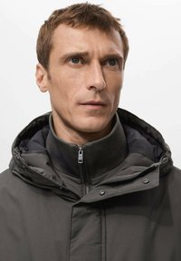 Giacca isolante verde scuro con zip frontale, colletto alto e cappuccio. Presenta un tessuto testurizzato e dettagli a pressione sul cancello.