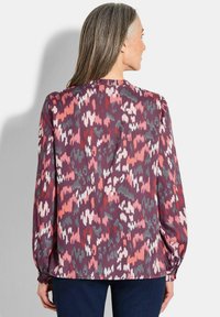 Langärmlige Bluse in den Farben Burgunderrot, Rosa und Grün mit einem abstrakten Muster. Hat geraffte Manschetten und einen runden Ausschnitt.