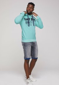 Felpa turchese con testo grafico e lacci, abbinata a pantaloni corti in denim grigi con risvolti blu e sneakers bianche.