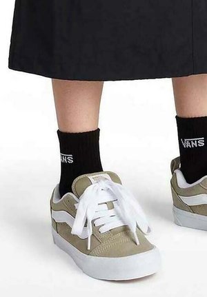 Zapatillas con parte superior de ante beige, rayas laterales de cuero blanco y cordones blancos. Combinadas con calcetas negras que tienen texto en blanco "VANS".
