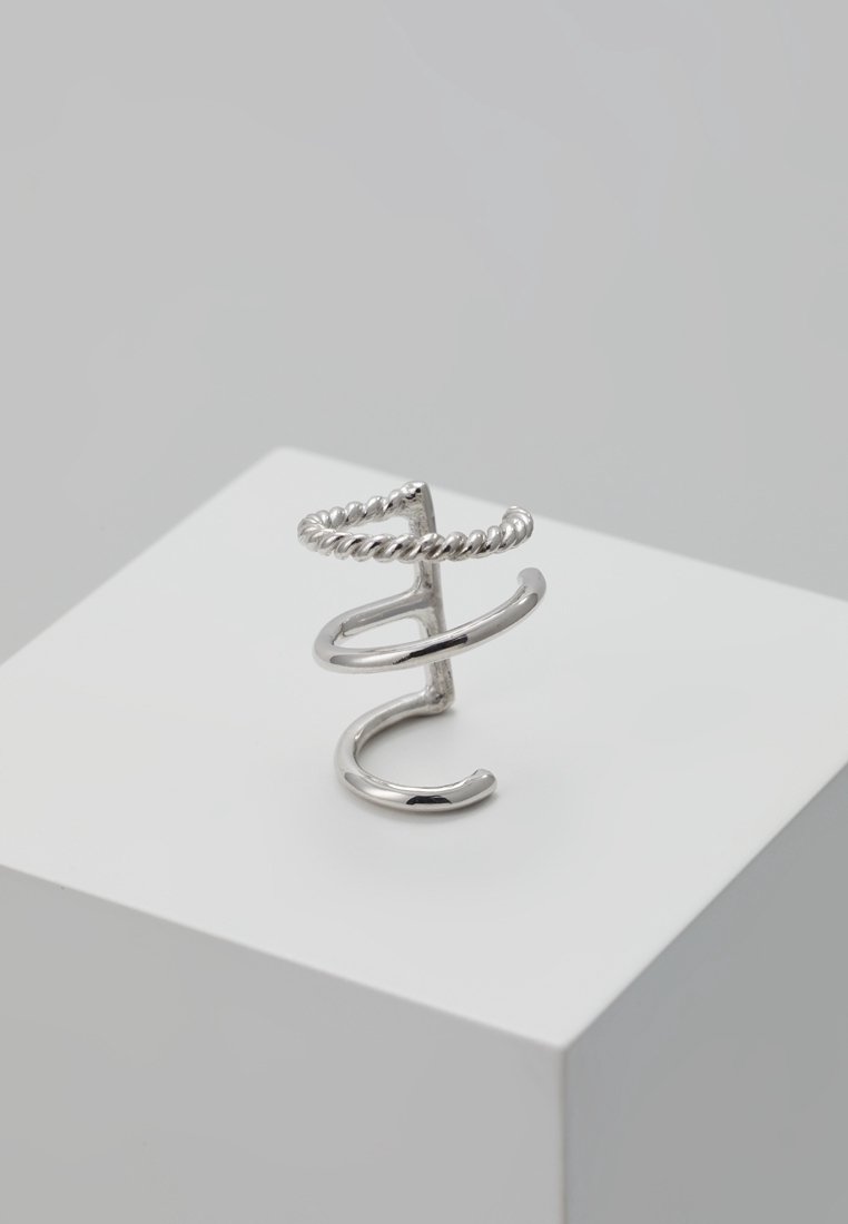 Boucle d'oreille triple bande ton argent avec bande supérieure torsadée présentée sur un socle géométrique blanc sur fond gris.