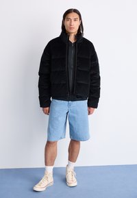 Giacca nera imbottita con design a quadri, colletto alto e zip frontale. Indossata con pantaloncini di jeans azzurri e sneaker beige alte.