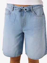 Ανοιχτό μπλε denim σορτς με δέσιμο στη μέση, μπροστινές τσέπες και κούμπωμα με κουμπί, φορεμένα με λευκό τοπ, κομμένα στη μέση.