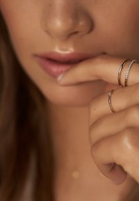 Trois bagues en or sur une main touchant le visage : une simple, une avec un petit diamant et une avec de petites pierres claires. Fond de peau lisse.