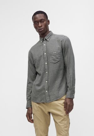 Homme portant une chemise à carreaux noir et blanc boutonnée et un pantalon cargo beige, debout devant un fond clair uni.