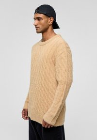 Beige kabelbrei sweater gemaakt van zachte, gestructureerde stof. Heeft een ronde halslijn, lange mouwen en een licht oversized pasvorm.