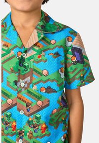 Camisa de manga corta con un colorido estampado gráfico de un paisaje en bloques, personajes y diversas texturas en verdes y azules.