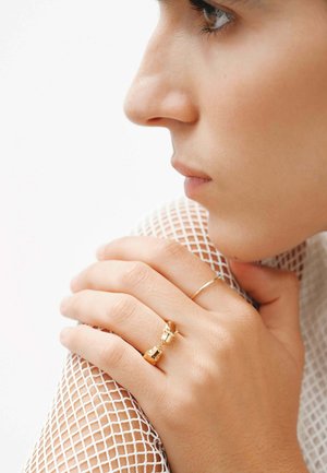 Anillos de oro con motivos de animales en los dedos, sobre una tela de malla blanca texturizada. Vista de perfil que resalta el tono de piel y los contornos suaves.