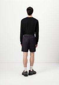 Chemise noire à manches longues, shorts sombres, tissu texturé, ceinture élastique, coutures visibles. Chaussures de sport grises avec des chaussettes blanches. Fond uni.