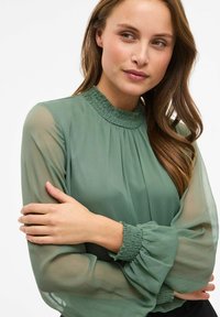 Grøn chiffonbluse med høj, smocket hals og lange, gennemsigtige ærmer med rynkede manchetter. Glat tekstur og flydende design.