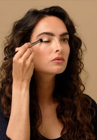 Applicazione del trucco in corso. Modella con lunghi capelli ricci e castani che utilizza un pennello da trucco nero sulla palpebra. Sfondo neutro.