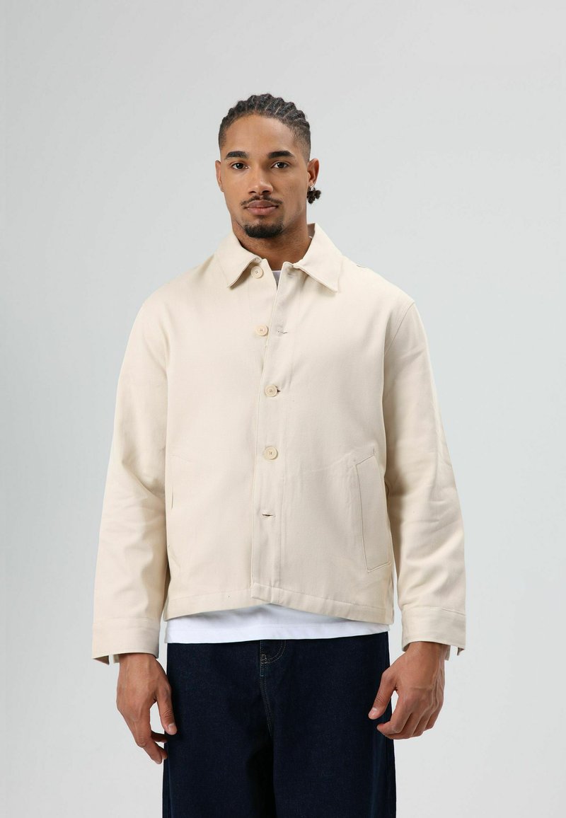 Beige canvasjack met een kraag, knoopsluiting aan de voorkant, twee zijzakken en lange mouwen. Gedragen over een wit shirt.
